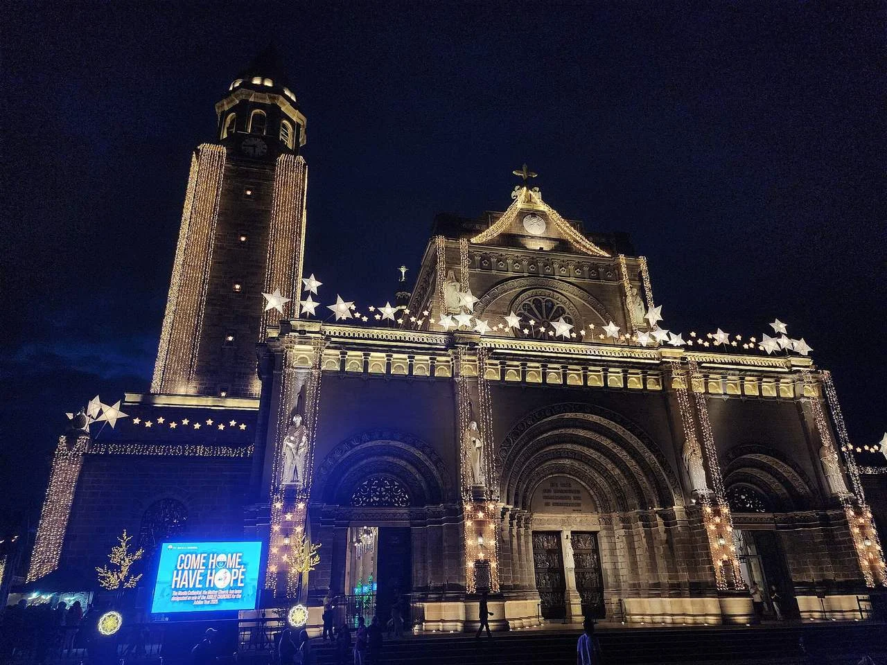 Manila Cathedral lit with Christmas lights and star lanterns before dawn Simbang Gabi, with a Come Home, Have Hope screen