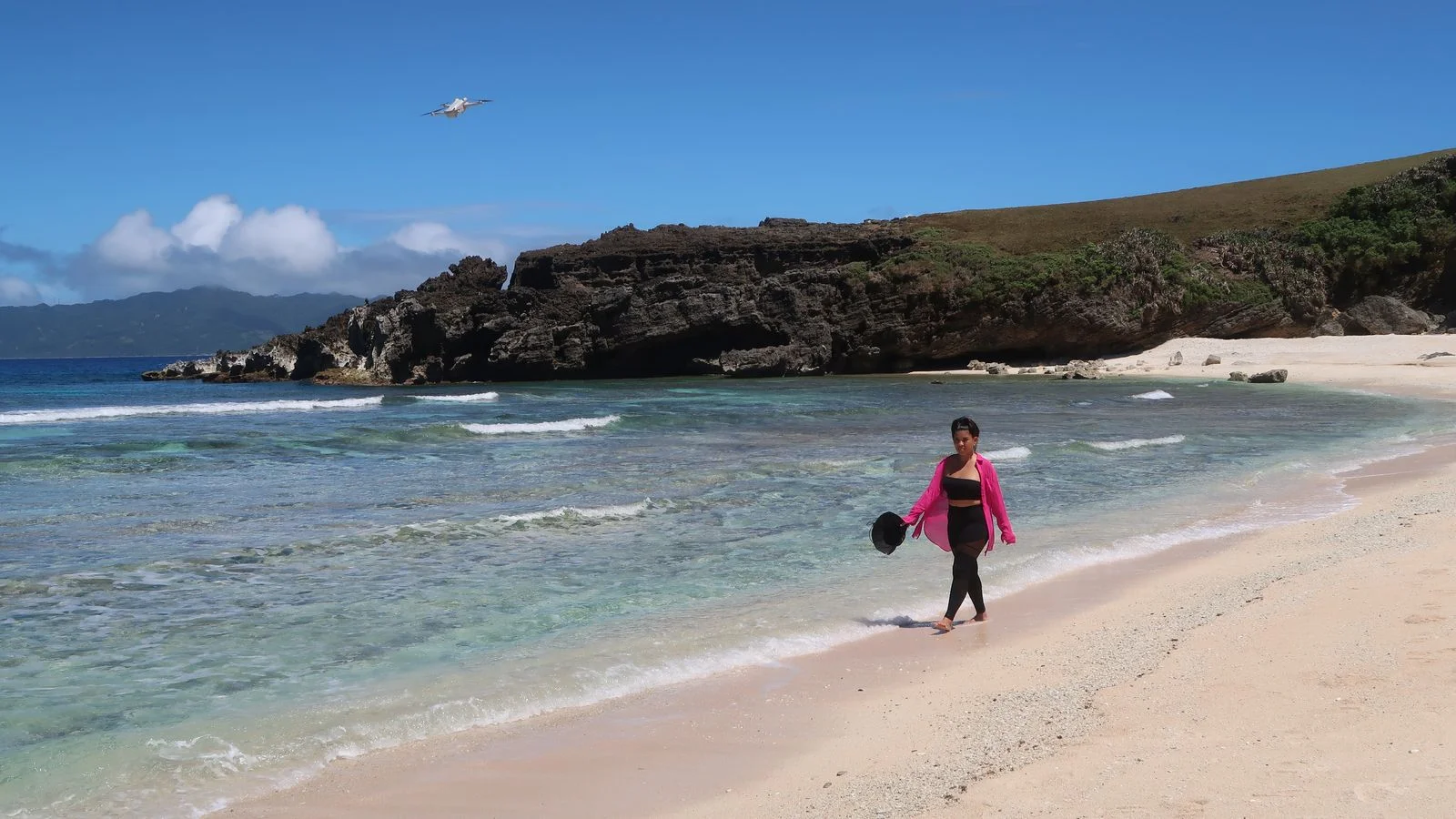 Woman walking along a white sand cove in Batanes with a rocky headland behind her and a small plane overhead